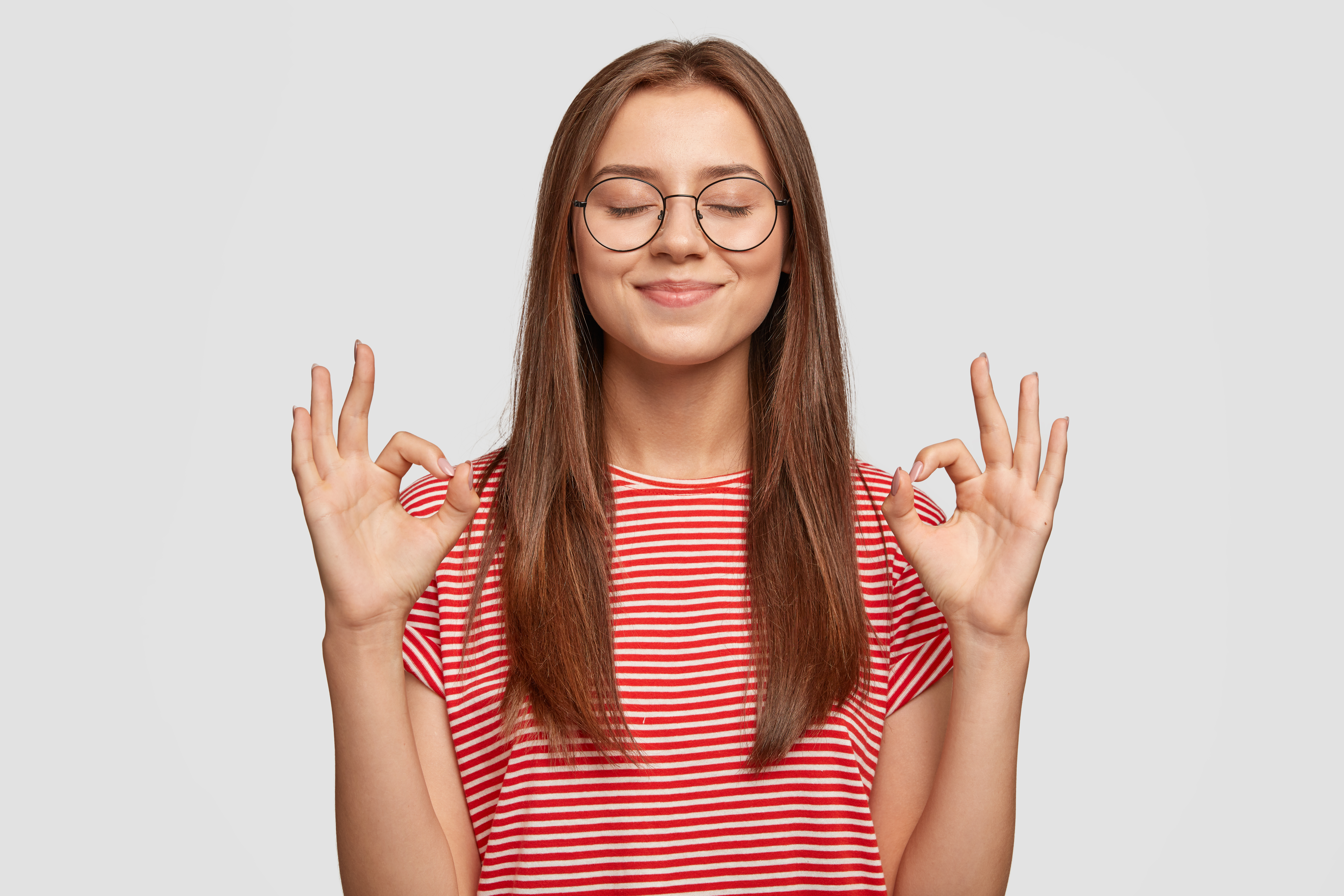 Pleased satisfied young female model makes zero gesture, wears transparent glasses, has long dark hair, dressed in casual striped t shirt, stands over white background. People, body language concept